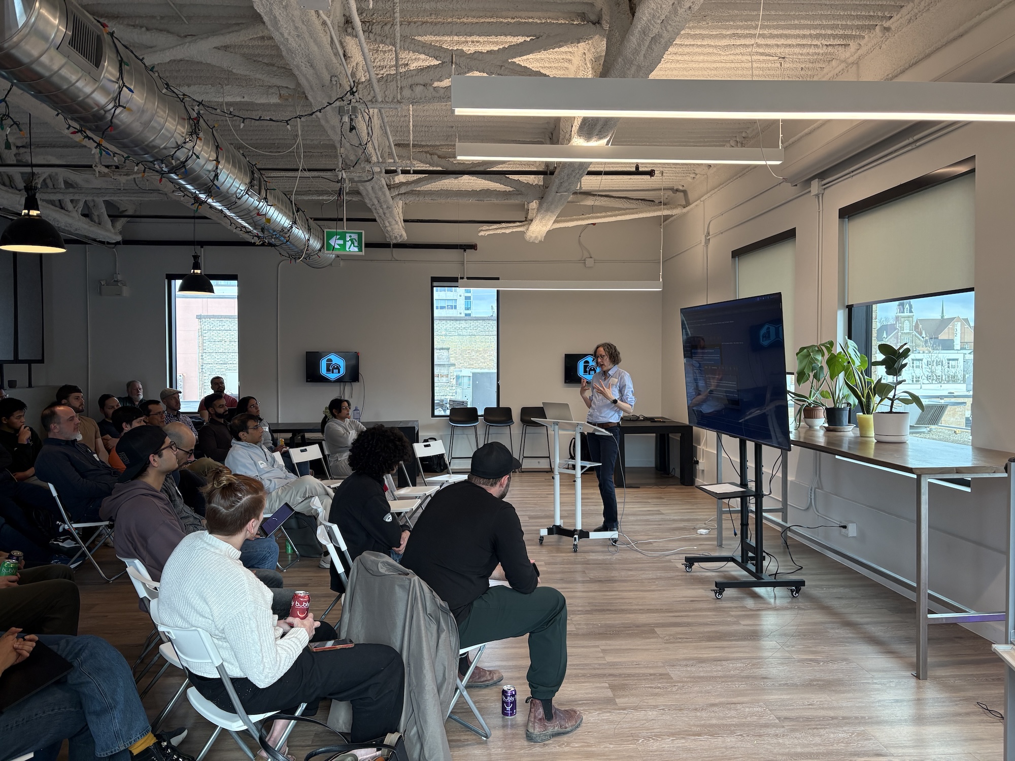 Presenter speaking beside a screen with the Claude logo to a seated audience at a Claude Waterloo meetup