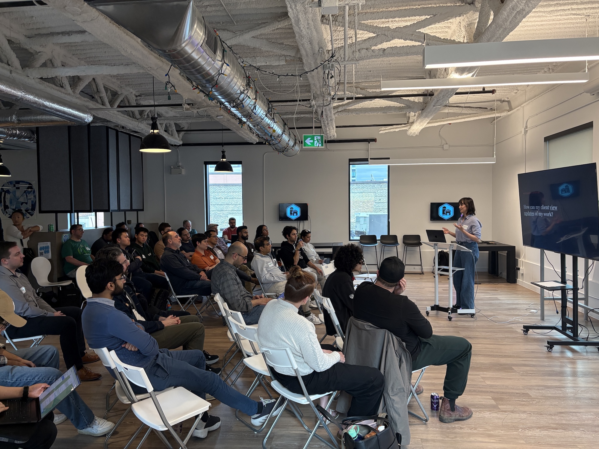 Presenter at a standing desk beside the big screen with a full seated audience in a bright loft at a Claude Waterloo meetup